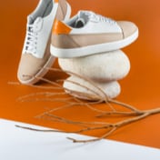 Beige and brown sneakers, one shoe standing with his side view on top of two small rocks. There is a small twig shown on the front.